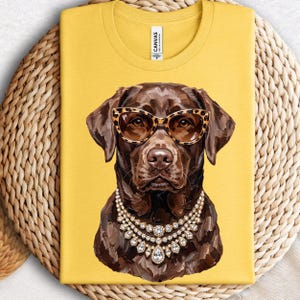 May include: A bright yellow t-shirt featuring a graphic illustration of a brown Labrador retriever wearing leopard print cat-eye sunglasses and a multi-layered crystal statement necklace. The shirt has a crew neckline and a white brand label.