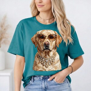 A woman with blonde hair wears a teal t-shirt featuring a graphic of a Labrador Retriever wearing leopard-print sunglasses and layered jeweled necklaces.