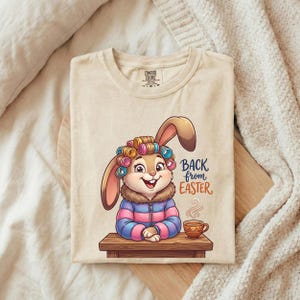 May include: Cream-colored t-shirt featuring a cartoon bunny with curlers, a jacket, and a cup of coffee. The text "BACK from EASTER" is printed on the right side of the bunny. The shirt is folded on a wooden surface.