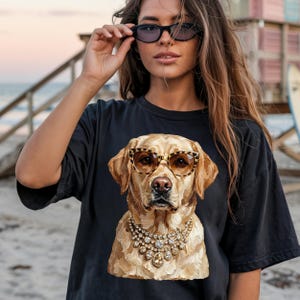 A person wearing a black t-shirt printed with a graphic of a glamorous Labrador Retriever in leopard print sunglasses and a diamond necklace stands on a beach at sunset, holding sunglasses up to her face.