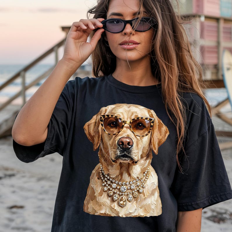 A person wearing a black t-shirt printed with a graphic of a glamorous Labrador Retriever in leopard print sunglasses and a diamond necklace stands on a beach at sunset, holding sunglasses up to her face.