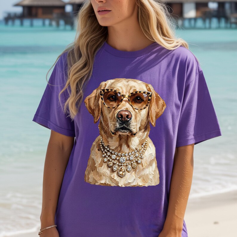 A person wears a purple t-shirt featuring an impasto graphic of a Labrador Retriever with leopard-print sunglasses and jeweled necklaces, with a beach and ocean in the background.