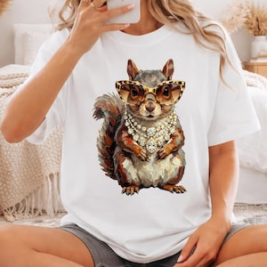 May include: A white t-shirt featuring a graphic print of a squirrel wearing leopard-print cat-eye sunglasses and a multi-layered gold and diamond-style necklace. The squirrel is rendered in a textured, painterly style with brown and gray fur tones.