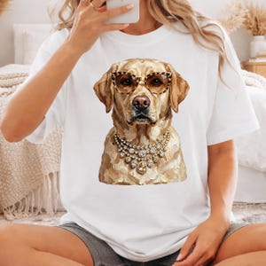 A person wearing a white t-shirt and grey shorts sits on the floor, showcasing a printed graphic of a golden retriever wearing leopard-print glasses and a crystal necklace.