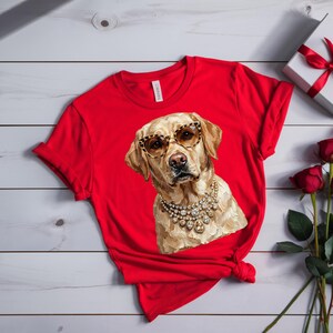 A bright red crewneck t-shirt lies on a white wooden surface next to a wrapped white gift box with a red ribbon and several red roses.