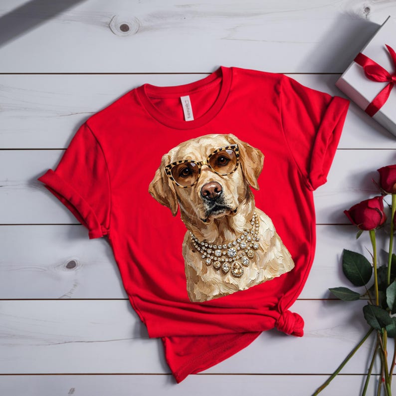 A bright red crewneck t-shirt lies on a white wooden surface next to a wrapped white gift box with a red ribbon and several red roses.