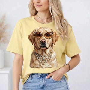 A woman wears a light yellow t-shirt printed with a golden Labrador Retriever wearing leopard-print glasses and heavy rhinestone necklaces.