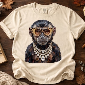 Puede incluir: Una camiseta de manga corta color crema con un estampado gráfico de un mono que lleva gafas con estampado de leopardo y un collar de cristales de varias capas. El mono está representado en un estilo pictórico con tonos de pelaje oscuro.