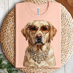 A peach-colored crewneck t-shirt rests on a round woven placemat, printed with an impasto illustration of a yellow Labrador Retriever wearing leopard-print glasses and diamond-encrusted necklaces.