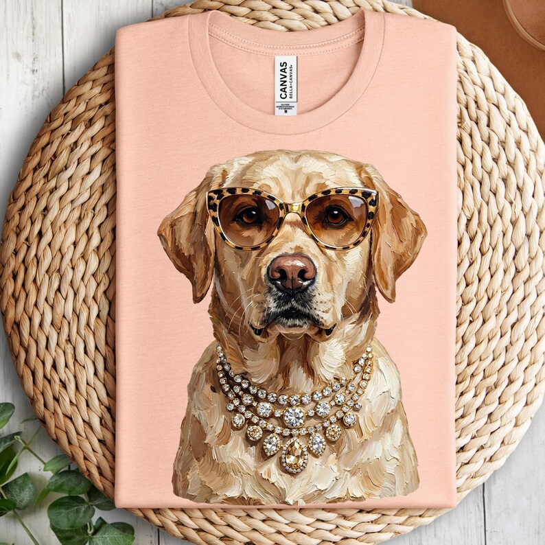A peach-colored crewneck t-shirt rests on a round woven placemat, printed with an impasto illustration of a yellow Labrador Retriever wearing leopard-print glasses and diamond-encrusted necklaces.