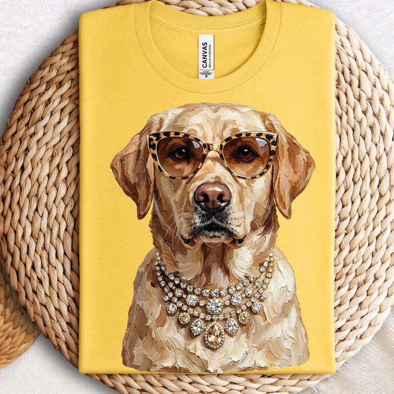 A mustard yellow t-shirt is folded on a round woven placemat, displaying a graphic of a yellow Labrador Retriever wearing leopard-print sunglasses and heavy jeweled necklaces.
