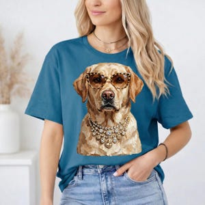 A blonde woman wearing a blue t-shirt printed with an impasto illustration of a Labrador Retriever in leopard print glasses and rhinestone necklaces poses indoors with her hand in her pocket.