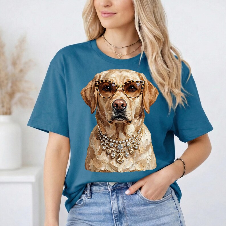 A blonde woman wearing a blue t-shirt printed with an impasto illustration of a Labrador Retriever in leopard print glasses and rhinestone necklaces poses indoors with her hand in her pocket.