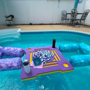 May include: A purple and yellow floating game table with a pile of small square tiles and a card in the center, flanked by a purple tie-dye pool float on the left and a blue tie-dye pool float on the right, with two drink bottles on the table.
