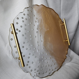 May include: A decorative resin tray with a clear, irregular shape. The tray features embedded pearl-like accents and gold-colored accents. It has two gold-colored handles. The tray is designed for serving or display.
