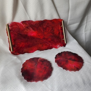 May include: A red resin tray with gold handles and two matching coasters. The tray and coasters have a marbled effect with shades of red and dark burgundy. The edges of the tray and coasters have a wavy, organic shape.