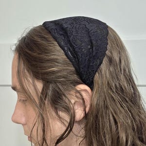 May include: A black lace headband. The headband is made of a delicate, floral-patterned lace. The hair is long and brown, with some strands falling over the face. The background is a plain white wall.