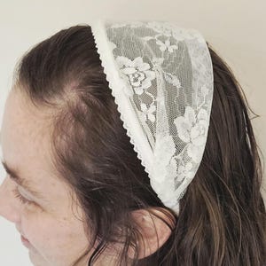 May include: A white lace headband featuring a floral design. The headband is crafted from delicate, see-through lace and has a scalloped edge. The headband is worn on the head.