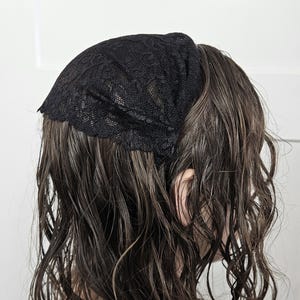 May include: A black lace head covering is worn on the head. The head covering is made of a delicate, open-weave lace fabric. The hair is dark brown and wavy. The background is a plain white wall.