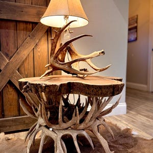 May include: A rustic side table with a unique design. The table features a natural wood slab top supported by a base crafted from deer antlers. A lampshade sits atop the table, adding a warm glow to the room. The lamp has a tan lampshade.