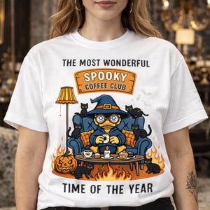 Könnte beinhalten: Weißes T-Shirt mit einem Halloween-Motiv. Das Design zeigt eine Cartoon-Ente mit Hexenhut, umgeben von schwarzen Katzen, einem Kürbis und Flammen. Der Text lautet "The Most Wonderful Spooky Coffee Club Time of the Year."