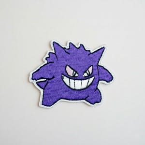 May include: A purple embroidered patch featuring a cartoon ghost with a wide grin and sharp teeth. The ghost has spiky features and pink accents around the eyes. The patch is outlined with white stitching.