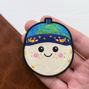May include: Embroidered patch featuring a smiling moon with rosy cheeks and a blue Earth-shaped hat. The patch has a black border and a small loop at the top. The moon has yellow freckles and a blue band with stars and crescent moons.