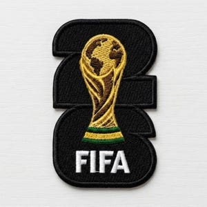 May include: A black embroidered patch featuring the number 2026 in a bold font. Centered over the numbers is a gold and green embroidered FIFA World Cup trophy icon. The word FIFA is stitched in white capital letters at the bottom of the patch.