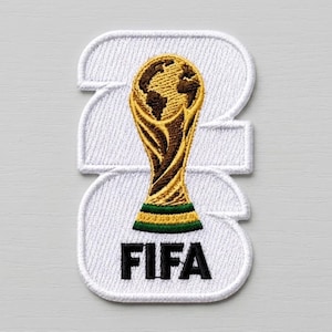 May include: A white embroidered patch featuring the number 2026 in a bold font. Centered over the numbers is a gold and brown embroidered trophy icon with a green base. The black text FIFA is embroidered at the bottom of the white patch design.
