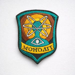 May include: Embroidered patch with a shield shape. The design features an atom symbol in white and yellow, set against a teal and brown background. The word "MOHOЛИТ" is displayed on a yellow banner at the bottom. The patch has a black border.