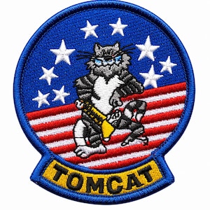 May include: A circular embroidered patch featuring a grey and white striped cat character holding a yellow object against a background of red and white horizontal stripes and white stars on a blue field, with the word TOMCAT in black on a yellow tab.