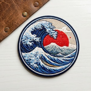 May include: Embroidered patch featuring a stylized depiction of a large ocean wave with white crests, a red sun, and a mountain range. The patch is circular with a dark blue border and is likely for decorative purposes.