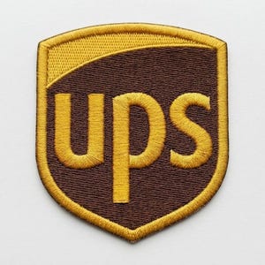 May include: A shield-shaped embroidered patch featuring the brown and yellow UPS logo. The design includes a yellow top section and a brown lower section with the letters UPS stitched in yellow thread, all enclosed by a yellow border.