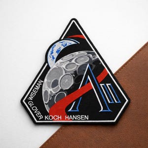 May include: A black, triangular patch featuring a stylized moon, Earth, and red and blue accents. The patch includes the names WISEMAN, GLOVER, KOCH, and HANSEN. The patch is on a white surface with a brown leather-like material in the background.