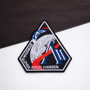 May include: Embroidered patch with a black, white, red, and blue design. The patch features a stylized moon, a red and blue striped element, and the names "WISEMAN", "GLOVER", "KOCH", and "HANSEN". The patch is in a triangular shape.