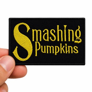 May include: A rectangular black embroidered patch featuring the yellow text Smashing Pumpkins. The patch has a thick black stitched border and a textured fabric surface with the lettering clearly displayed in a stylized serif font.