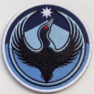 May include: A circular embroidered patch featuring a black bird with a red eye, wings spread wide, centered against a light blue background. A white eight-pointed star sits above the bird, all enclosed within a dark blue border and a white outer edge.