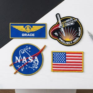 May include: Four embroidered patches with space and patriotic themes. One patch features the NASA logo, another displays the American flag, and a third reads "GRACE" with wings. The fourth patch says "PROJECT HAIL MARY".