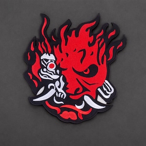 May include: An embroidered patch featuring a stylized red and black demon mask design with flame-like edges. The graphic includes white accents, sharp teeth, and a prominent eye, all outlined in black thread against a dark gray background.