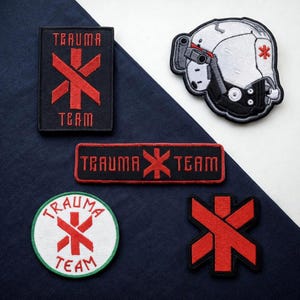 May include: Collection of embroidered patches featuring the words "TRAUMA TEAM" and a red medical symbol. Includes a patch shaped like a futuristic helmet with a medical symbol. Patches are black, red, white, and green.
