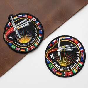 May include: Two circular embroidered patches with the text "PROJECT HAIL MARY". The patches feature a rocket design with multiple flags around the border. The background is black with red, white, and yellow accents. The patches are on a brown leather surface.