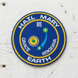May include: A circular patch with a blue background and yellow border. The text "HAIL MARY" and "EARTH" are in white, with additional text in other languages. The patch features a stylized design with a blue arc and green plus signs.