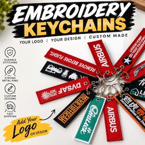 May include: A collection of rectangular fabric keychains featuring embroidered text and logos in various colors including red, black, and teal. Each keychain includes a metal ring attached through a reinforced eyelet, displaying custom designs.