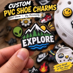May include: A custom PVC shoe charm featuring a mountain landscape with green pine trees, a yellow sun, and blue water waves, with the text EXPLORE in white letters. The charm is held against a background of various other colorful shoe charms.