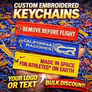 Może przedstawiać: Niestandardowe haftowane breloczki do kluczy w kolorze czerwonym, niebieskim i żółtym. Breloczki mają tekst "REMOVE BEFORE FLIGHT", "CALIFORNIA RADOMES" i "MADE IN SPACE FOR ATHLETES ON EARTH". Na zdjęciu widnieje również napis "YOUR LOGO OR TEXT" i "BULK DISCOUNT!"