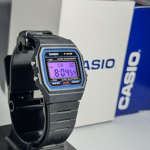 May include: Black Casio F-91W digital watch with a blue and purple display, resting on a black stand. The watch face displays the time and day. The watch is next to a white and blue Casio box.