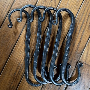 May include: Set of five handcrafted, dark gray metal S-hooks. Each hook features a twisted central shaft and curled ends. The hooks are arranged on a wooden surface, showcasing their rustic design.