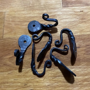 May include: A collection of six hand-forged, black metal hooks. Each hook features a unique design with twisted stems and curled ends. The hooks vary in shape and size, with mounting holes for easy installation. The hooks are displayed on a wooden surface.