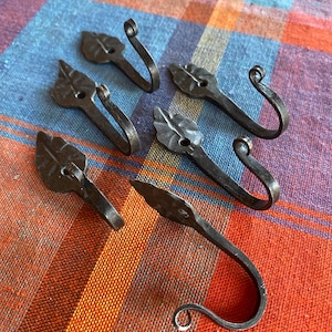 May include: Six handcrafted, dark metal leaf-shaped hooks. Each hook features a leaf design and a curved end for hanging items. The hooks are arranged on a colorful, patterned fabric background.