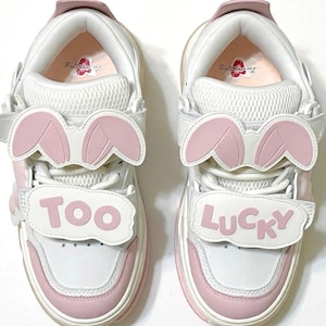 May include: A pair of white and light pink sneakers featuring bunny ear attachments on the laces and velcro straps across the front. The left shoe displays the word TOO and the right shoe displays the word LUCKY in pink lettering on white patches.